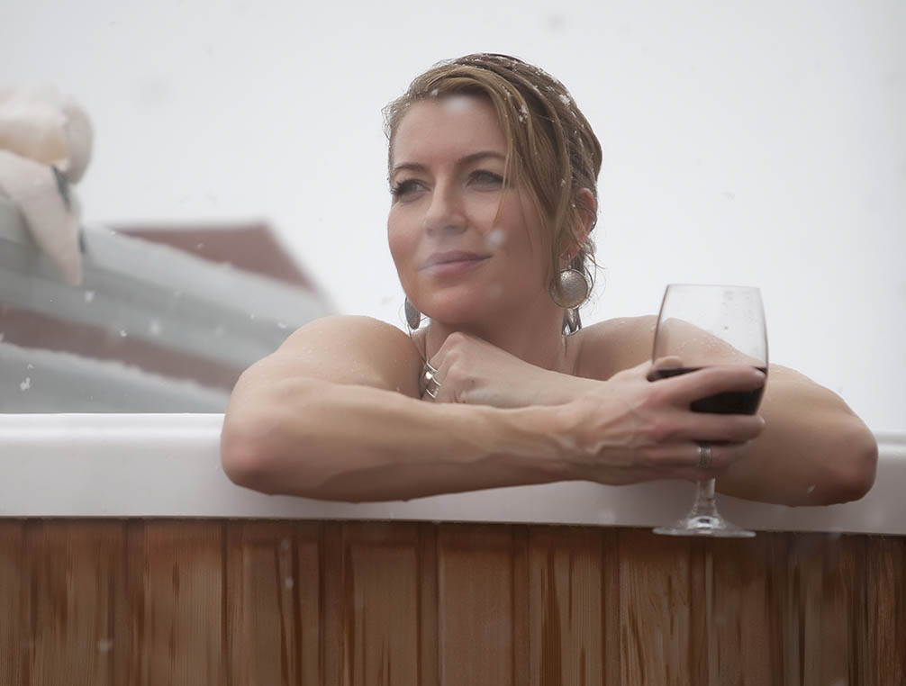 Woman in hot tub with glass of wine leaning against edge looking at snow